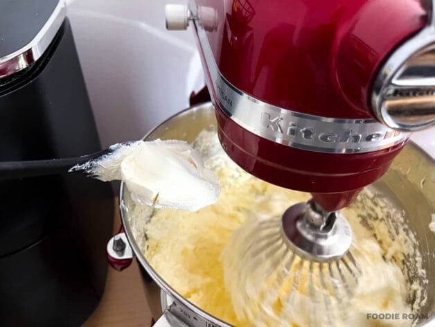 A spoonful of mascarpone cheese being added to the buttercream mixture in a stand mixer.