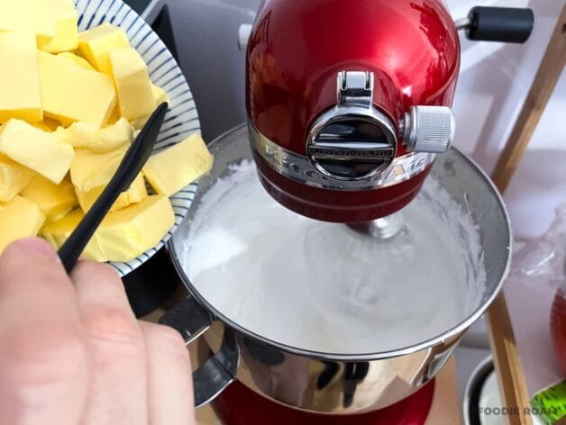 Softened butter cubes being added to whipped meringue in a stand mixer.