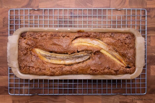 A loaf of banana bread topped with halved caramelized bananas, cooling in a parchment-lined pan on a wire rack.