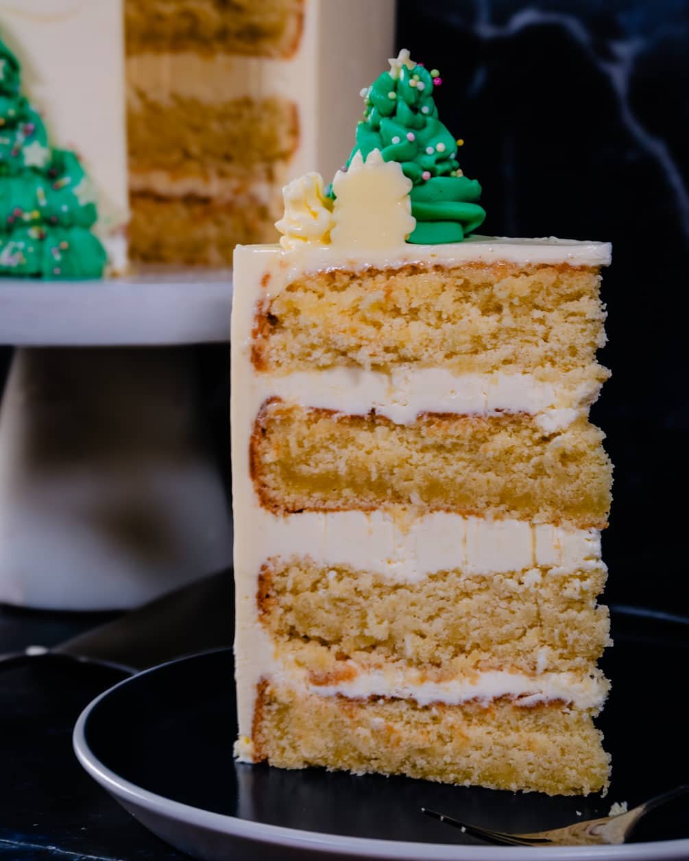 A slice of festive Coconut Almond Cake with layers of almond mascarpone buttercream, decorated with green buttercream Christmas trees.