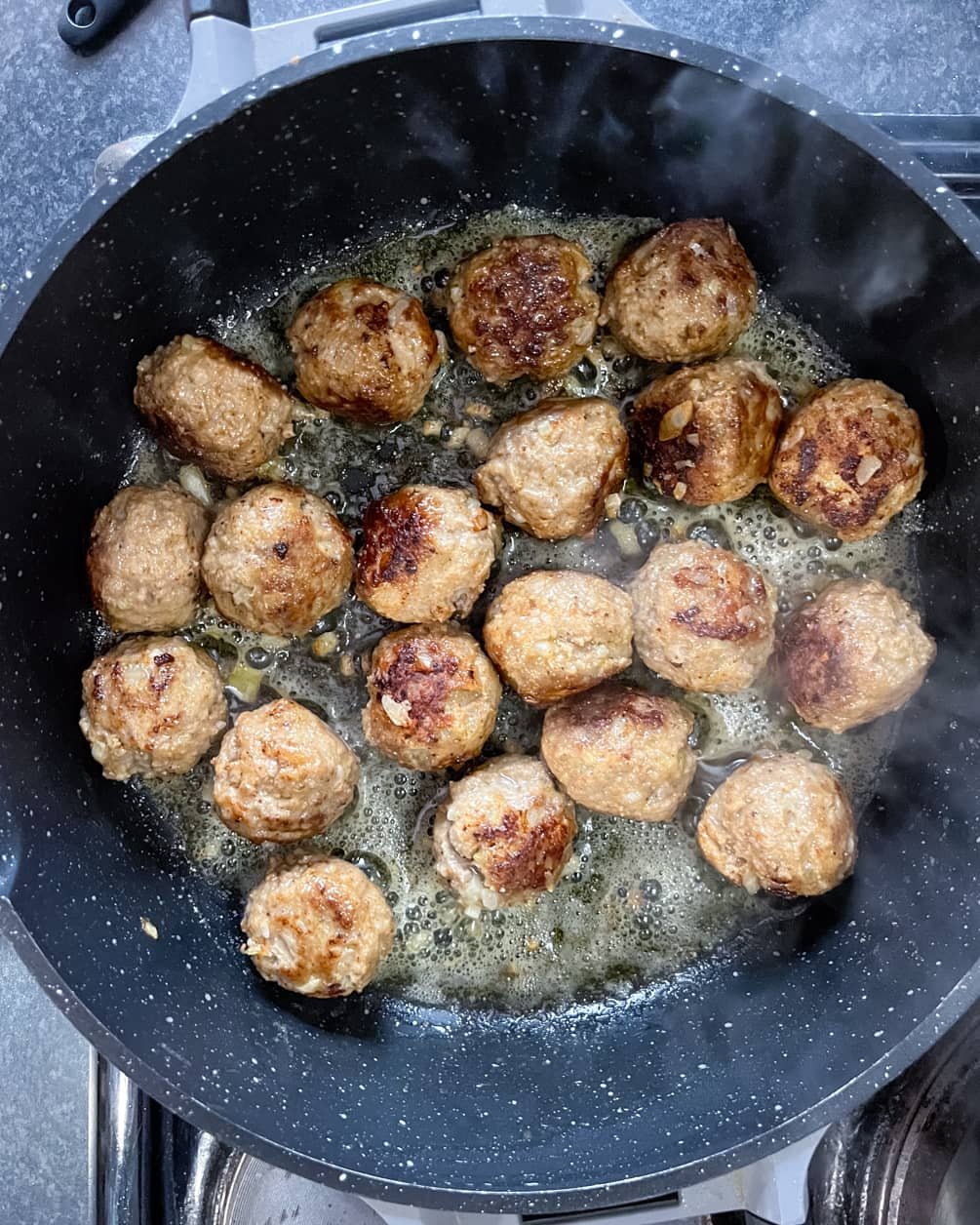 Golden-brown meatballs frying in bubbling butter in a non-stick pan, releasing steam as they cook.