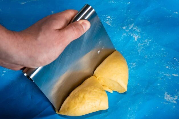 A hand holding a dough scraper, dividing freshly kneaded pasta dough into smaller portions.