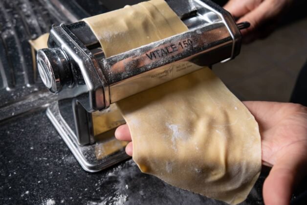 Fresh pasta dough being rolled through a stainless steel pasta machine to create thin, even sheets.