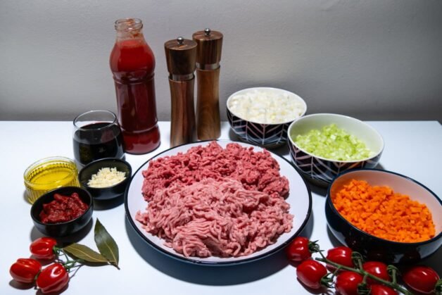 A neatly arranged display of ingredients for Dalmatian-style meat ragu, including ground pork and beef, chopped vegetables, passata, tomato paste, red wine, olive oil, garlic, and spices.