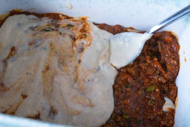 A spoon spreading creamy béchamel sauce over a layer of rich meat ragu in a baking dish.
