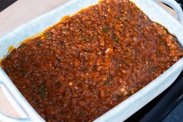 A layer of rich meat ragu spread evenly over fresh pasta sheets in a baking dish.