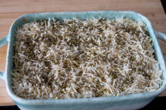 A baking dish filled with lasagna, topped with shredded mozzarella, Parmesan cheese, and Italian herb mix.