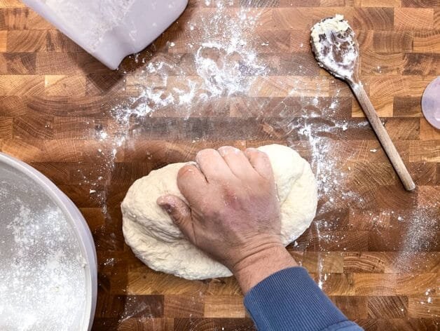 A hand kneading soft flatbread dough on a floured wooden surface with a wooden spoon and flour container nearby.