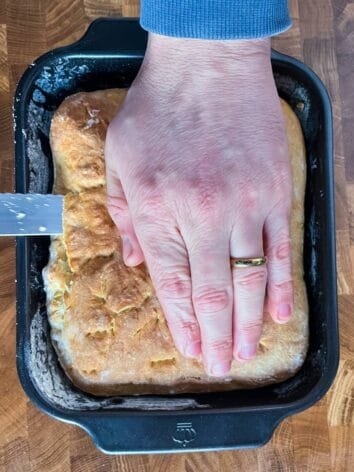 A hand holding down baked flatbread in a black baking pan while slicing off the top crust.
