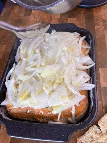 Sliced onions being drizzled with hot oil over flatbread in a black baking pan.