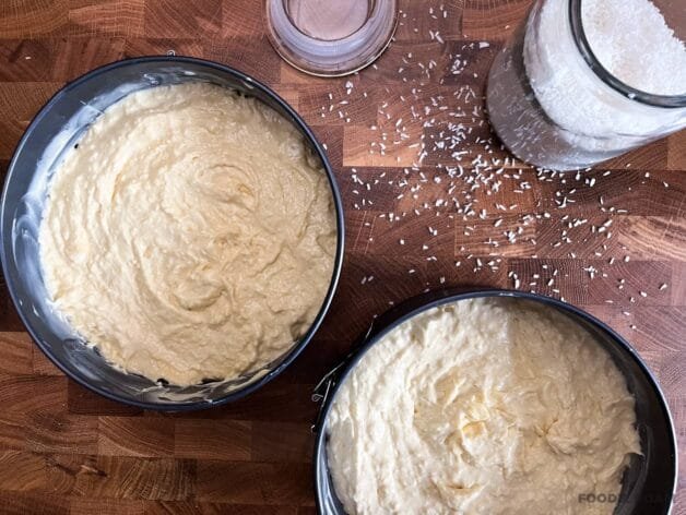 Cake batter evenly divided into two round baking pans, ready for the oven, with desiccated coconut scattered on the surface.
