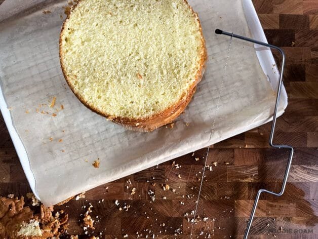 A leveled cake sponge with the caramelized top removed using a cake leveler, sitting on parchment paper with crumbs scattered around.