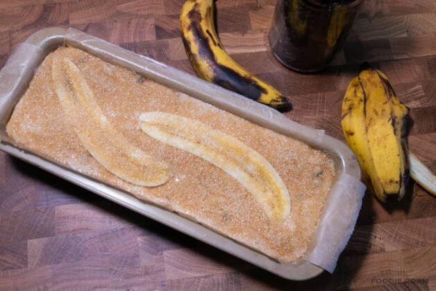 A loaf pan filled with banana bread batter, topped with halved bananas and sprinkled with demerara sugar.