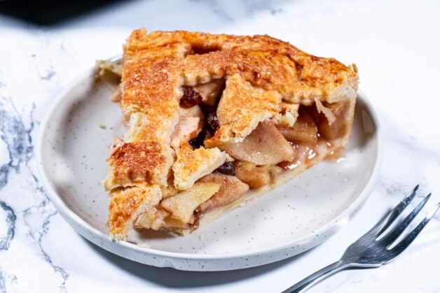 A slice of apple pie with a flaky crust, filled with spiced apples and raisins, served on a plate.