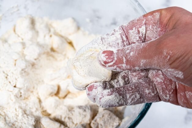 A hand flattening a butter cube coated in flour to create flaky layers for the pie crust.