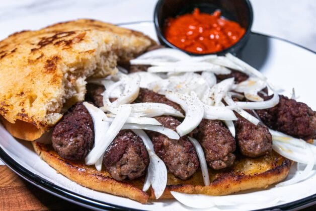 Open-faced cevapcici topped with sliced onions, served with a side of ajvar.
