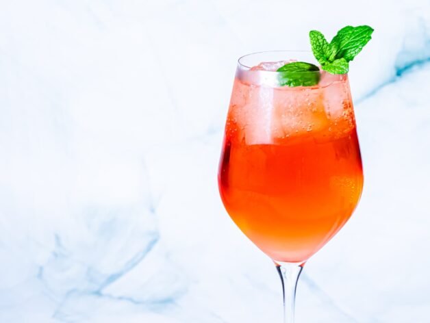 A Citrus Hennessy Spritz in a tall glass, with ice and mint garnish, placed on a light, textured surface.
