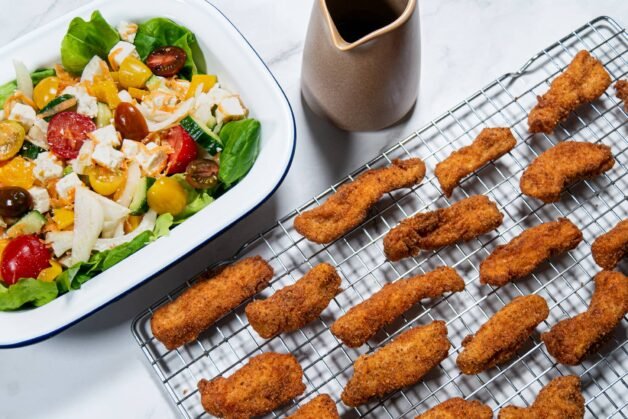 Crispy chicken strips on a cooling rack next to a colorful salad bowl with basil dressing.