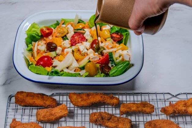 Hand pouring basil dressing onto a colorful salad with crispy chicken strips nearby.