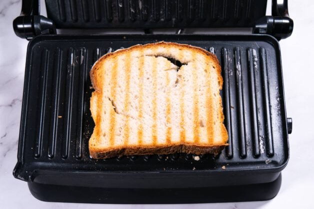 A slice of toasted bread on a grill press with golden-brown grill marks.