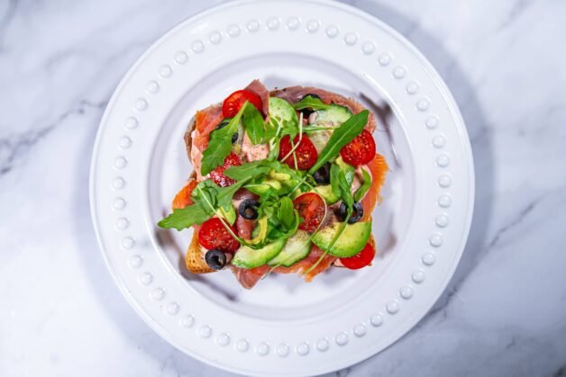 A fully assembled smoked salmon and avocado toast with fresh veggies and arugula on a white plate.