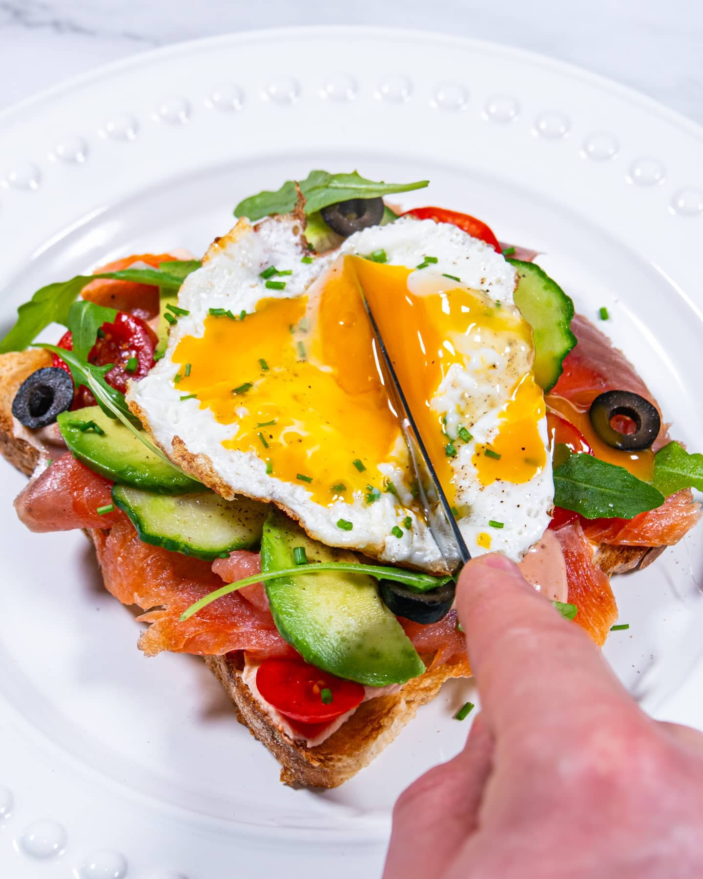 A knife cutting into the yolk of a sunny-side-up egg on smoked salmon and avocado toast, with layers of fresh veggies and arugula.