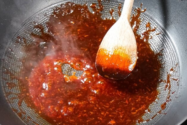 A wooden spoon stirring thick, glossy Yangnyeom sauce in a skillet, with steam rising.