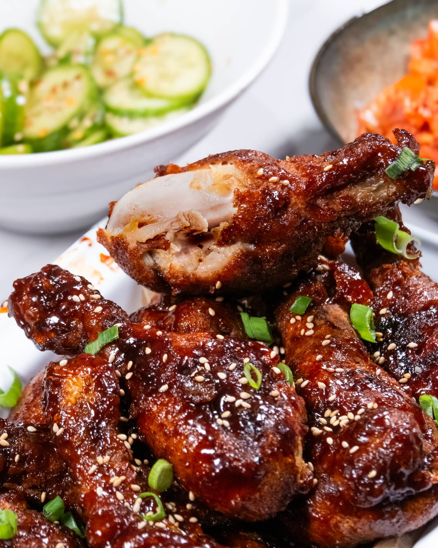Glazed Korean fried chicken drumsticks garnished with sesame seeds and chopped green onions, served with cucumber salad and kimchi.