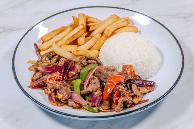 A plated serving of Lomo Saltado with beef, stir-fried vegetables, French fries, and a side of white rice.