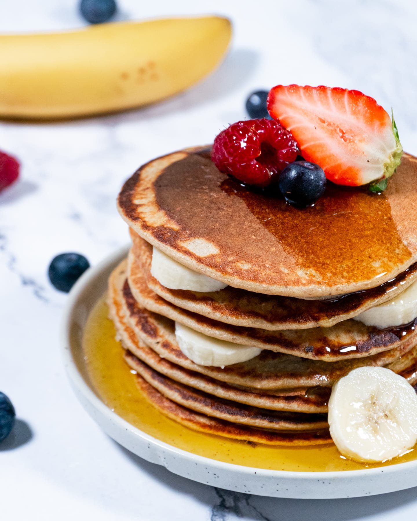 Stack of oatmeal pancakes topped with fresh fruit and maple syrup on a marble surface.