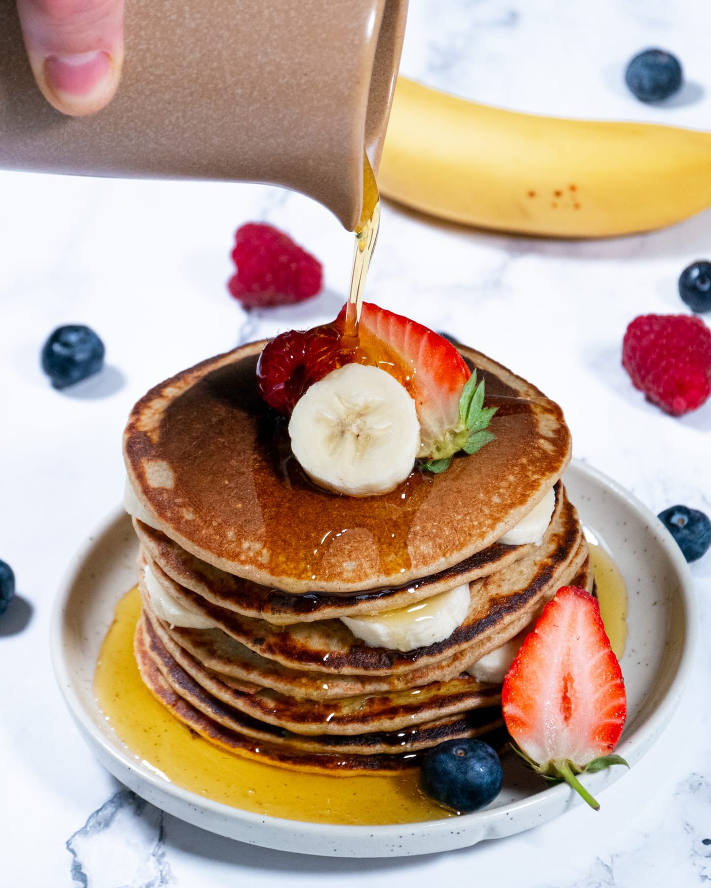 Syrup being poured over oatmeal pancakes stacked with banana slices and fresh berries.