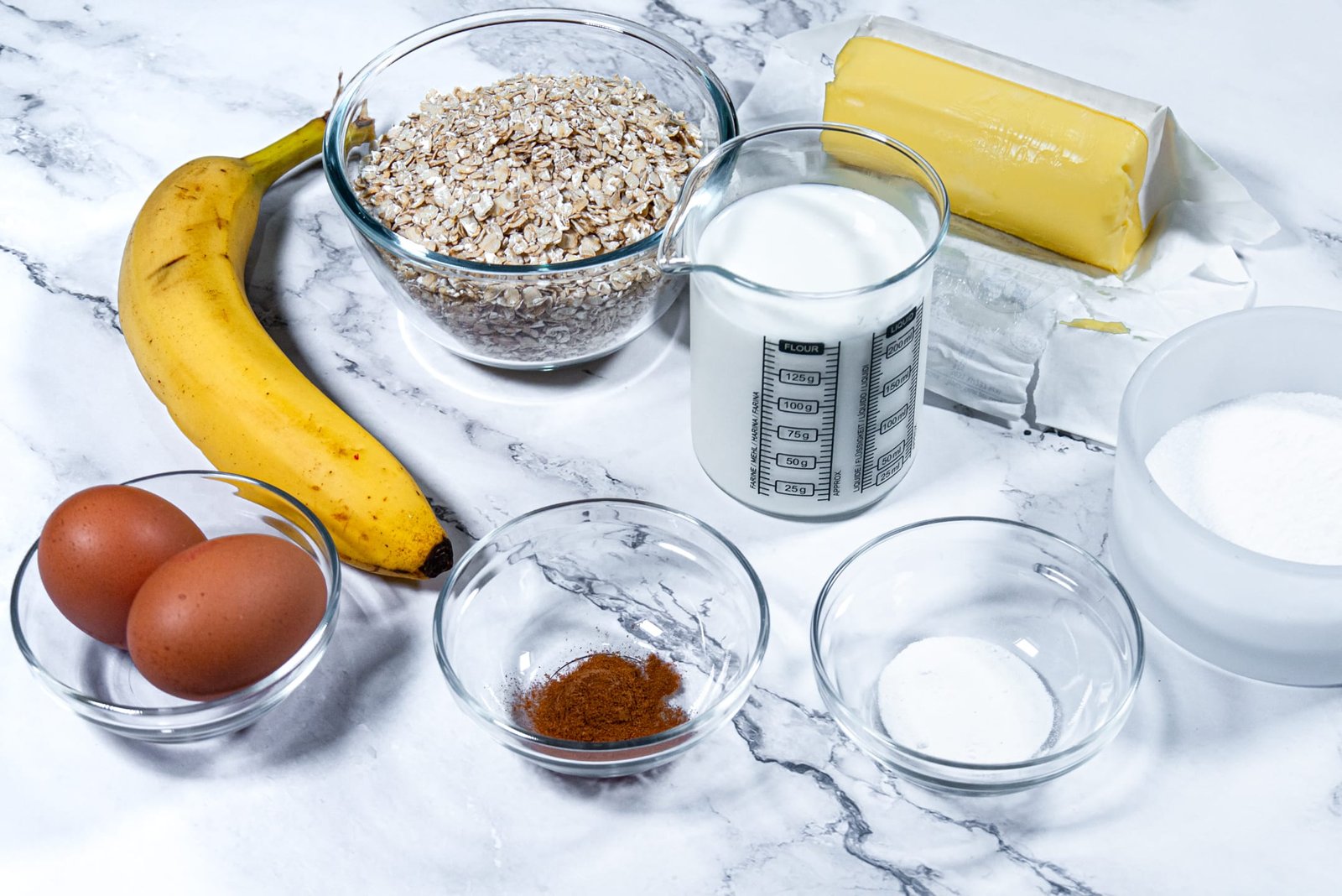 Ingredients for oatmeal pancakes on a marble surface, including oats, banana, milk, eggs, butter, baking powder, and cinnamon.