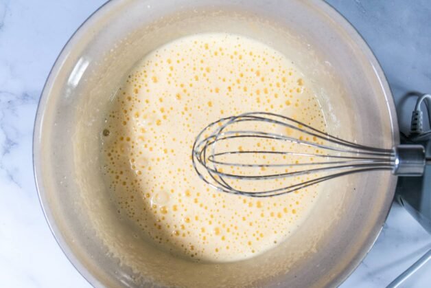 Light and frothy egg and sugar mixture in a mixing bowl, with a whisk resting on the side.