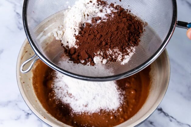 Cocoa powder and all-purpose flour being sifted into a chocolate mixture using a fine mesh sieve.