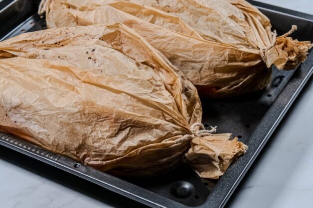 Two tightly wrapped Sea Bass en Papillote parcels tied with kitchen twine on a baking tray.