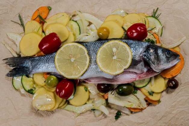 Whole sea bass on a bed of sliced vegetables with lemon slices on top, ready to be wrapped in parchment paper.