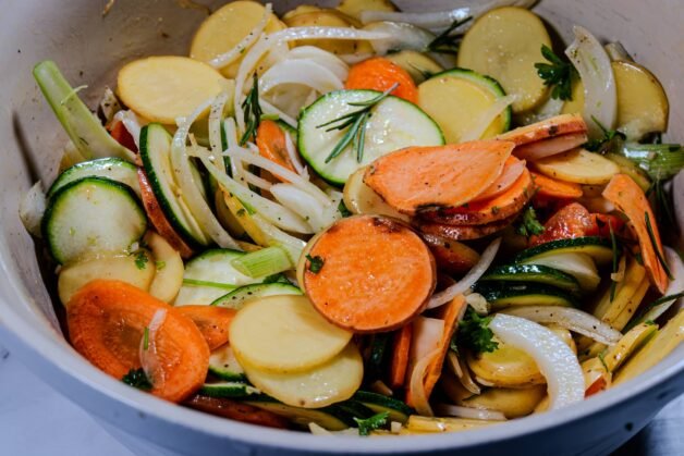 Sliced potatoes, zucchini, sweet potatoes, carrots, and onions mixed with rosemary and olive oil in a bowl.