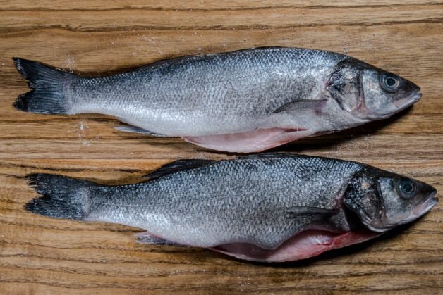 Two whole raw sea bass on a wooden cutting board, sprinkled with salt.