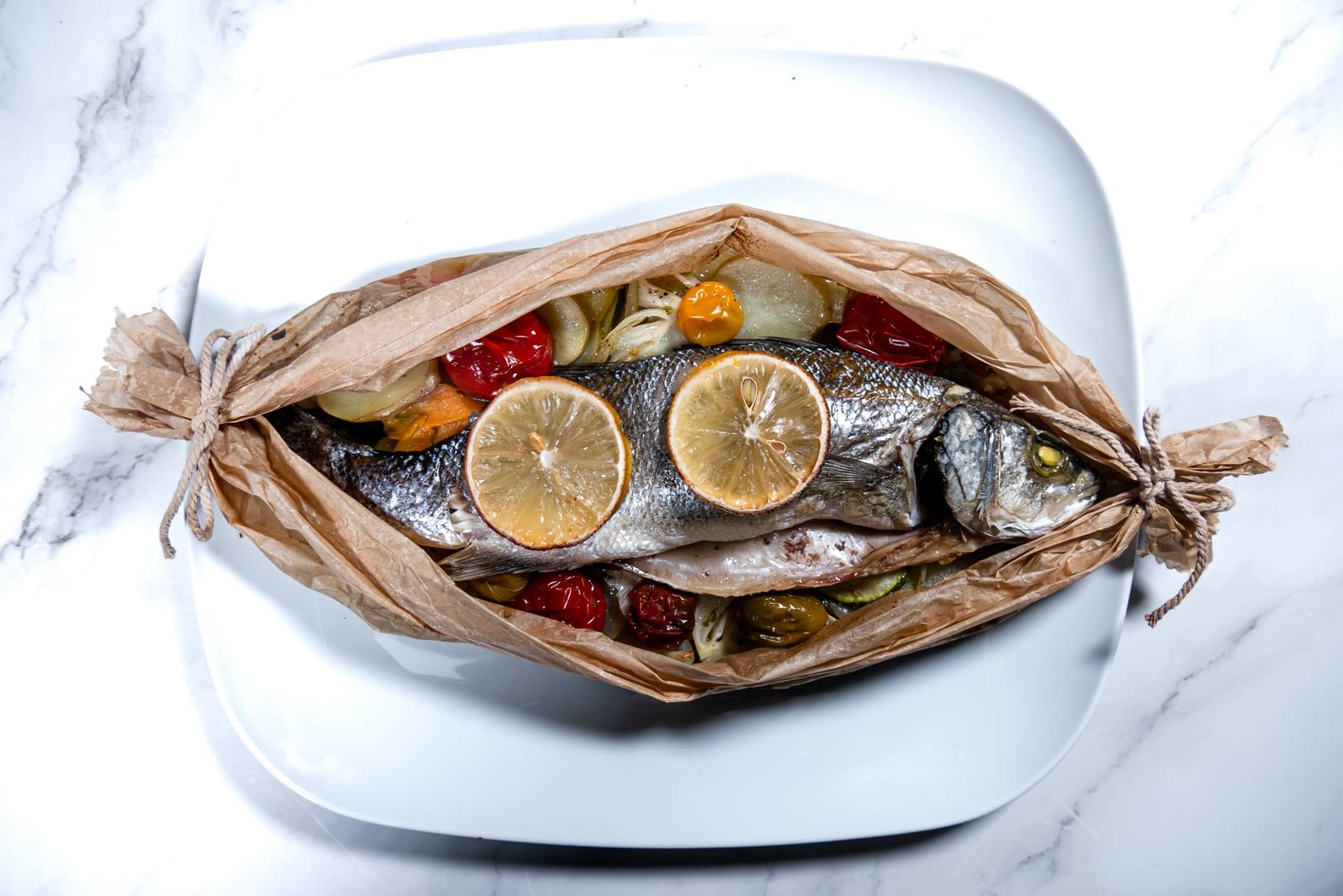 Sea Bass en Papillote with lemon slices, cherry tomatoes, and vegetables in parchment paper on a white plate.