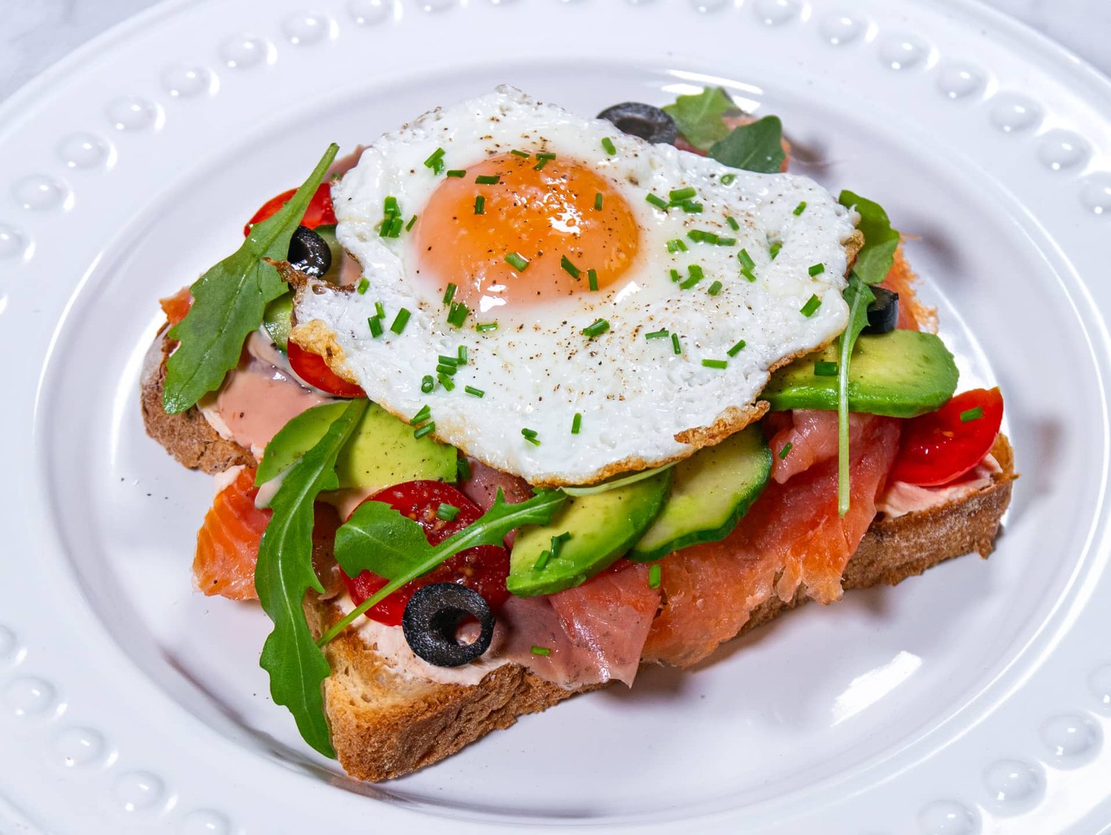 Smoked salmon and avocado toast topped with a sunny-side-up egg, garnished with fresh chives on a white plate.