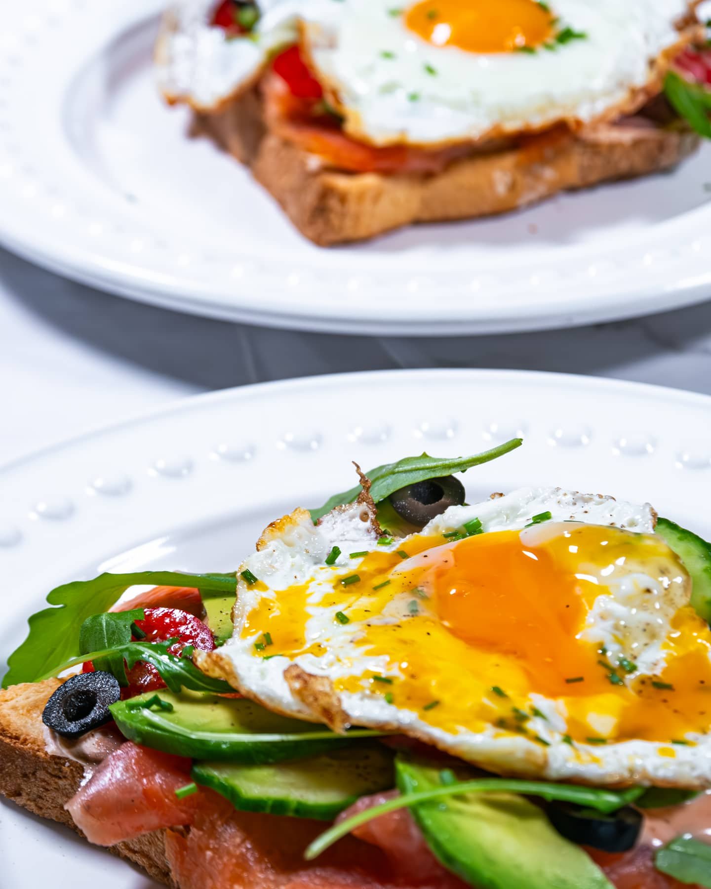 Two plates of smoked salmon and avocado toast topped with sunny-side-up eggs, garnished with fresh chives.