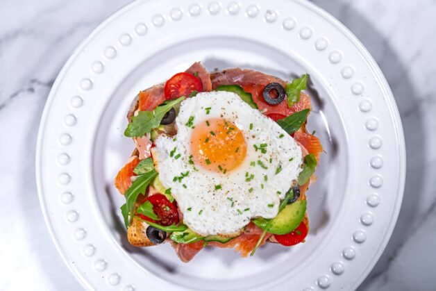 Top-down view of smoked salmon and avocado toast topped with a sunny-side-up egg, surrounded by vibrant veggies and garnished with fresh chives.