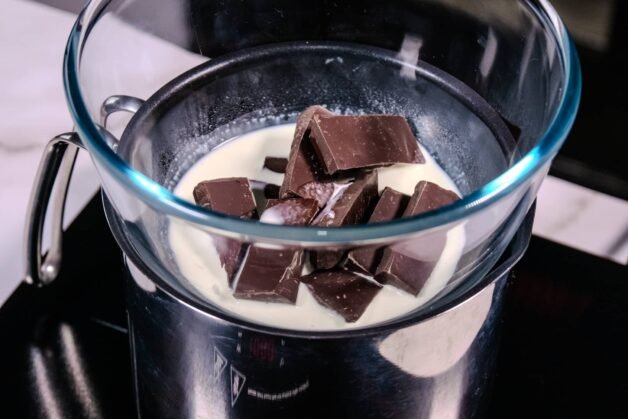Dark chocolate pieces melting with heavy cream in a glass bowl over a double boiler.