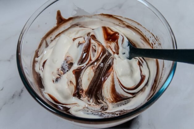 Whipped cream being folded into melted chocolate in a glass bowl with a spatula.