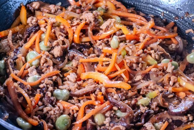A close-up of stir-fried ground pork with shiitake mushrooms, carrots, bell peppers, and broad beans in a wok.