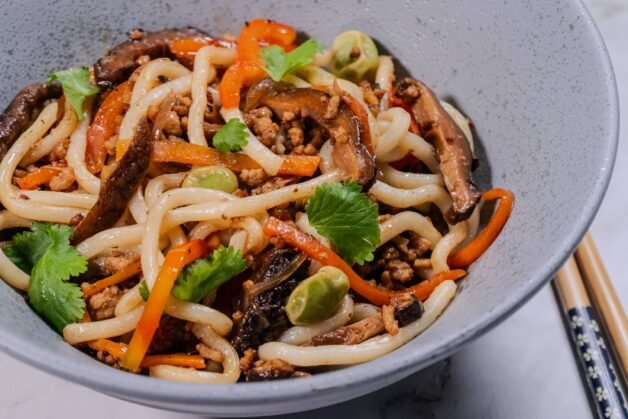 A close-up of udon noodle stir-fry with ground pork, shiitake mushrooms, bell peppers, and fresh cilantro in a gray bowl, with chopsticks on the side.