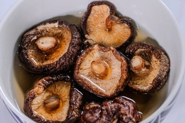 A close-up of dried shiitake mushrooms soaking in water in a white bowl, showing their rich texture and earthy tones.