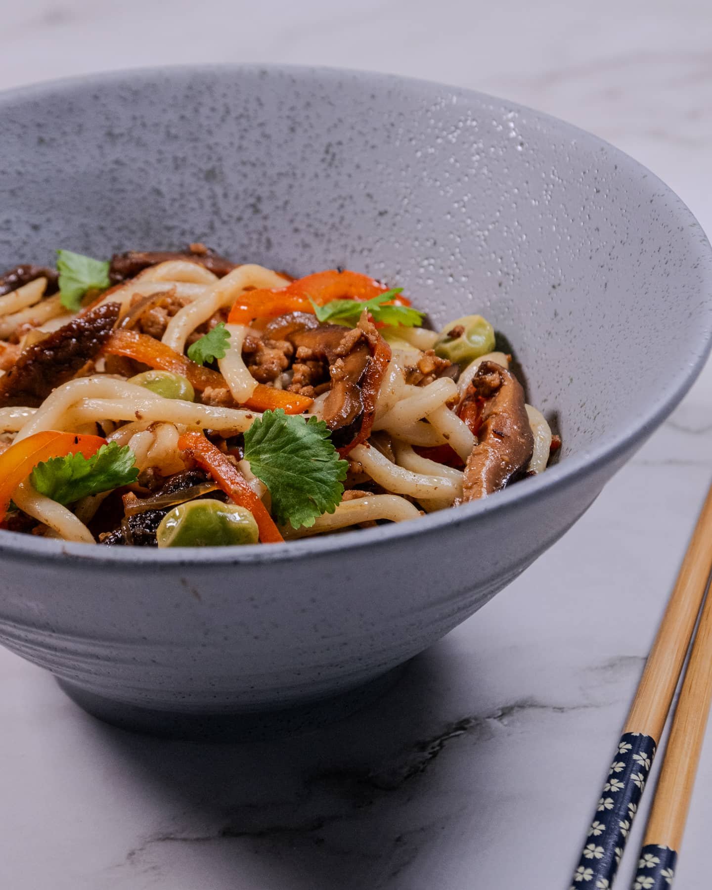 A close-up of a bowl of udon noodle stir-fry with ground pork, shiitake mushrooms, red bell pepper, and fresh cilantro garnish, served in a textured gray bowl with chopsticks on the side.