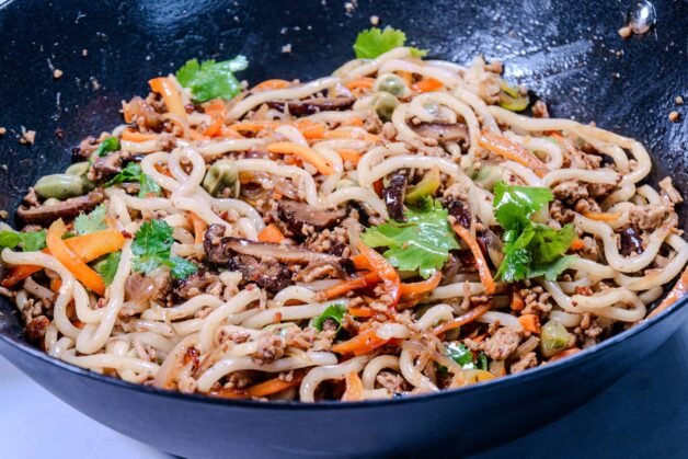 A wok filled with udon noodles stir-fried with ground pork, shiitake mushrooms, vegetables, and garnished with fresh cilantro.