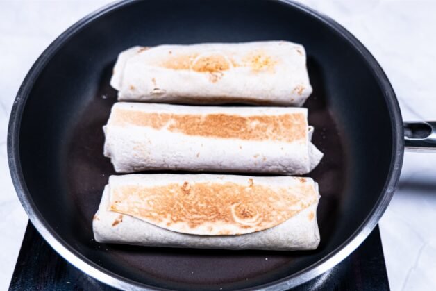 Three beef burritos being toasted in a skillet, developing a crispy, golden-brown crust.